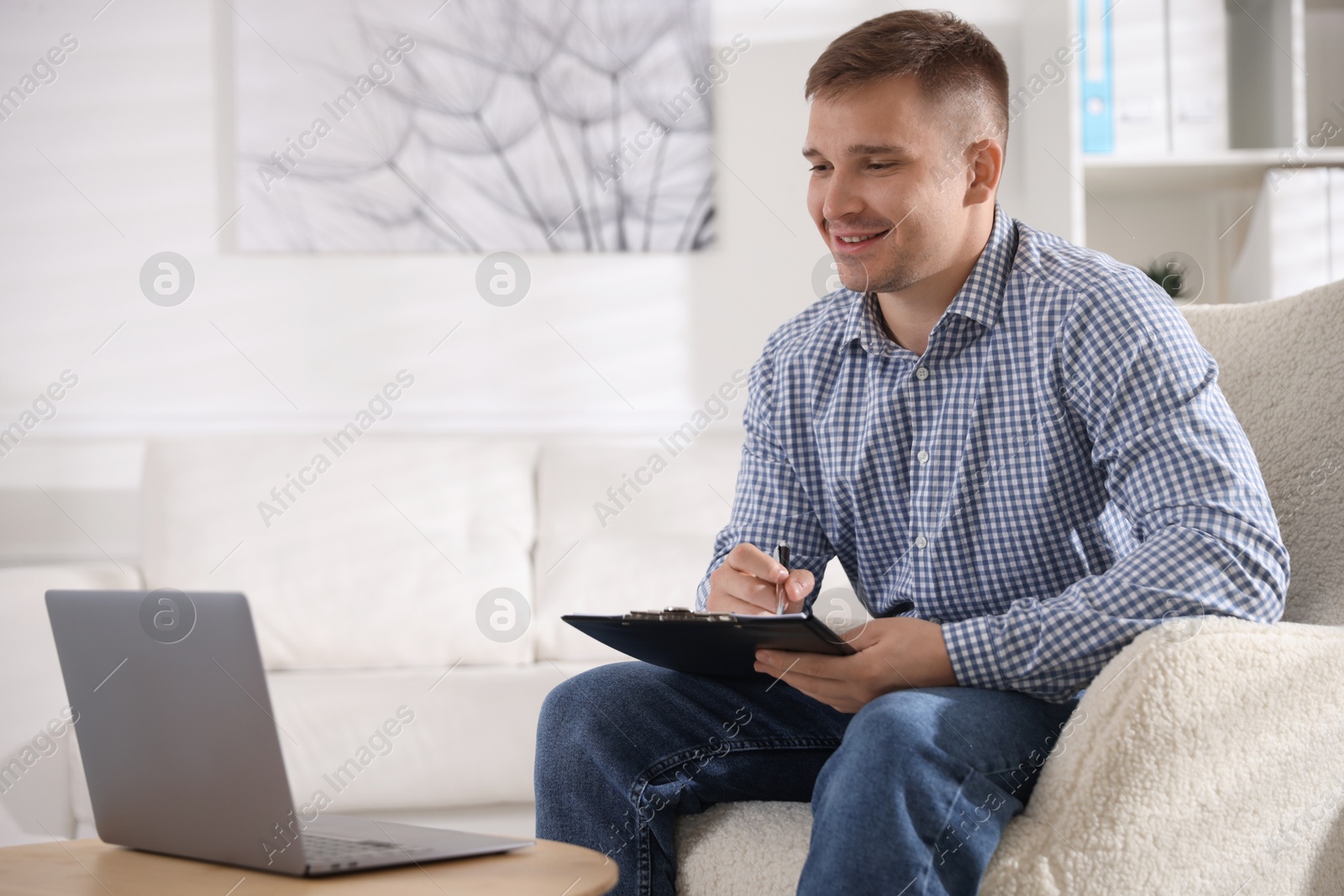 Smiling psychologist with clipboard during online session by laptop in office Photo of Smiling psychologist with clipboard during online session by laptop in office