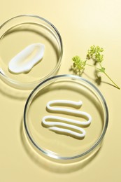 Photo of Different cosmetic products in Petri dishes and floral decor on pale yellow background, flat lay