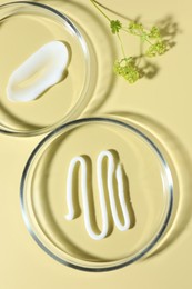 Photo of Different cosmetic products in Petri dishes and floral decor on pale yellow background, flat lay