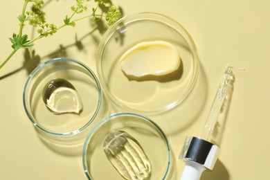 Photo of Different cosmetic products in Petri dishes, dropper and floral decor on pale yellow background, flat lay