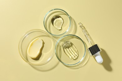 Photo of Different cosmetic products in Petri dishes and dropper on pale yellow background, flat lay