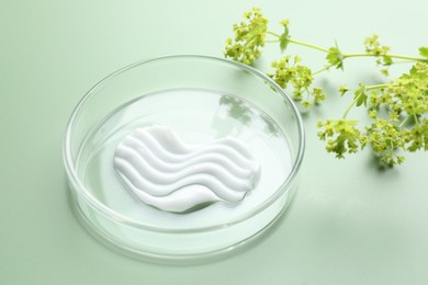 Photo of Cosmetic product in Petri dish and floral decor on green background, closeup