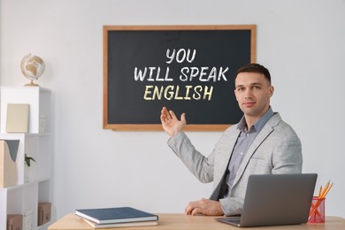 Image of Teacher pointing at text You Will Speak English on black chalkboard in classroom. Language course
