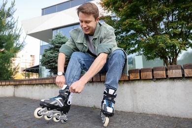 Photo of Happy young man putting on roller skates outdoors, low angle view