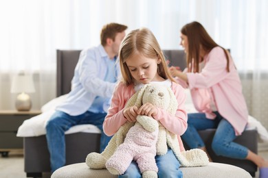 Photo of Upset little girl with bunny and her parents fighting at home, selective focus