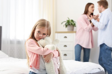 Photo of Upset little girl with bunny and her parents fighting at home, selective focus