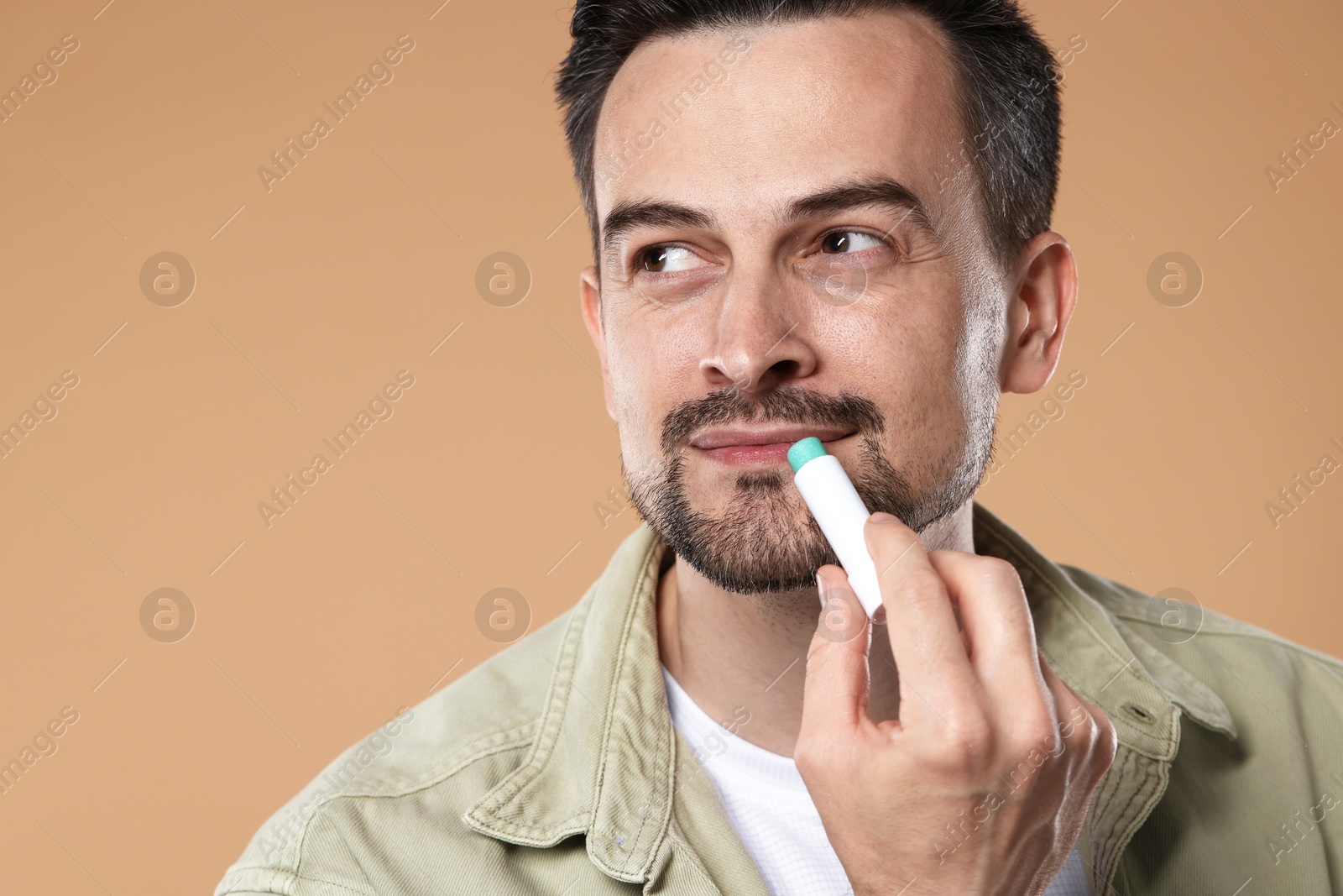 Photo of Handsome man applying lip balm onto his lips on light brown background