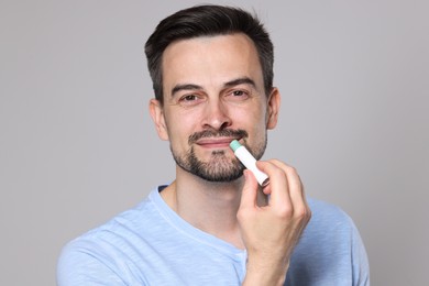 Photo of Handsome man applying lip balm onto his lips on grey background