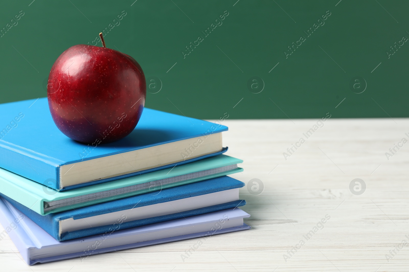 Different notebooks and red apple on white wooden table against blackboard, closeup. Space for text Photo of Different notebooks and red apple on white wooden table against blackboard, closeup. Space for text