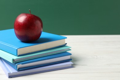 Photo of Different notebooks and red apple on white wooden table against blackboard, closeup. Space for text