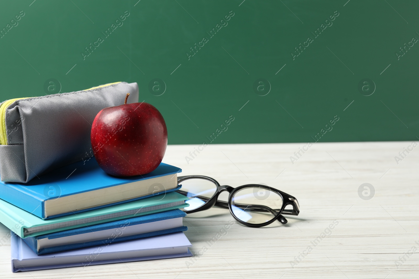 Different notebooks, pencil case, glasses and red apple on white wooden table against blackboard, closeup. Space for text Photo of Different notebooks, pencil case, glasses and red apple on white wooden table against blackboard, closeup. Space for text