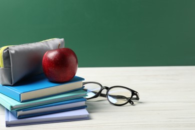 Photo of Different notebooks, pencil case, glasses and red apple on white wooden table against blackboard, closeup. Space for text