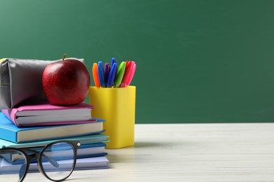 Photo of Different school stationery, glasses and red apple on white wooden table against blackboard, closeup. Space for text