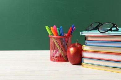 Photo of Different school stationery, glasses and red apple on white wooden table against blackboard, space for text