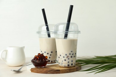 Photo of Tasty milk bubble tea in plastic cups, tapioca pearls and green leaves on beige table