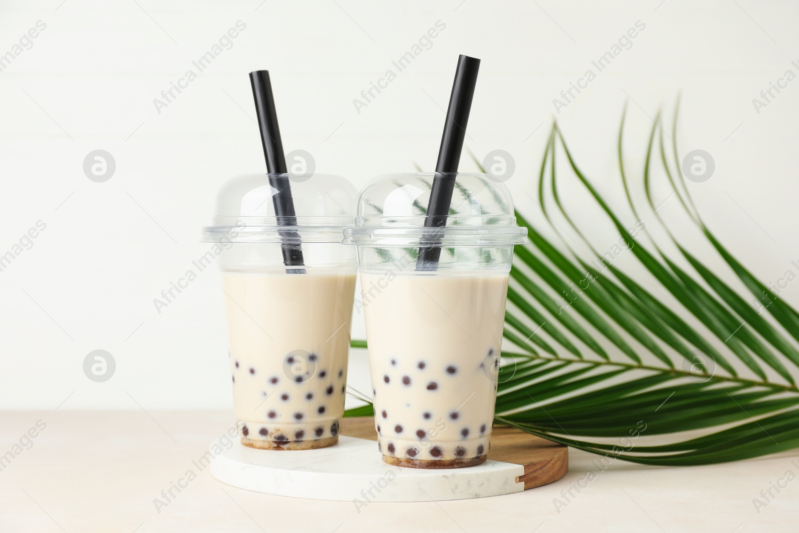Photo of Tasty milk bubble tea in plastic cups and green leaves on beige table