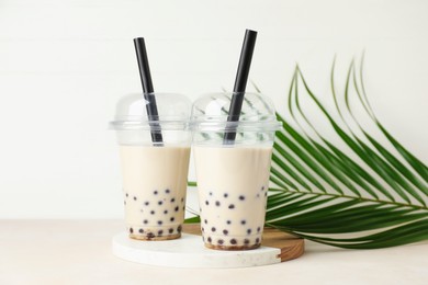 Photo of Tasty milk bubble tea in plastic cups and green leaves on beige table