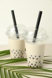 Photo of Tasty milk bubble tea in plastic cups and green leaves on beige table, closeup