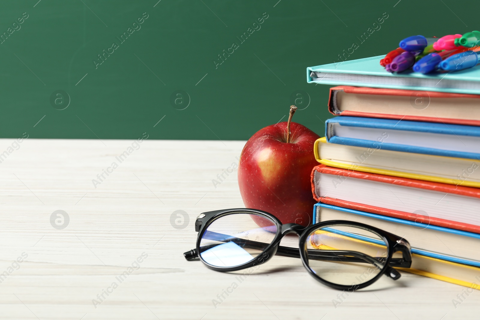 Different school stationery, glasses and red apple on white wooden table against blackboard, closeup. Space for text Photo of Different school stationery, glasses and red apple on white wooden table against blackboard, closeup. Space for text