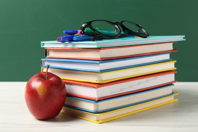 Photo of Different school stationery, glasses and red apple on white wooden table against blackboard, closeup