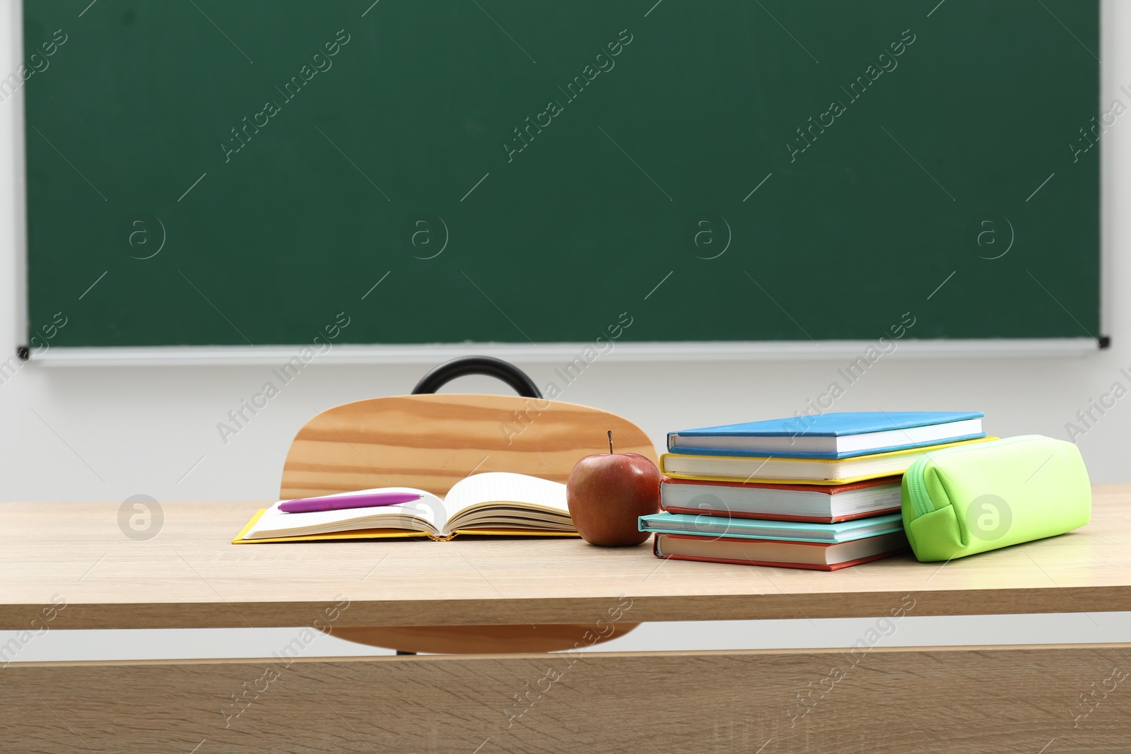 Different school stationery and apple on wooden table near blackboard indoors Photo of Different school stationery and apple on wooden table near blackboard indoors