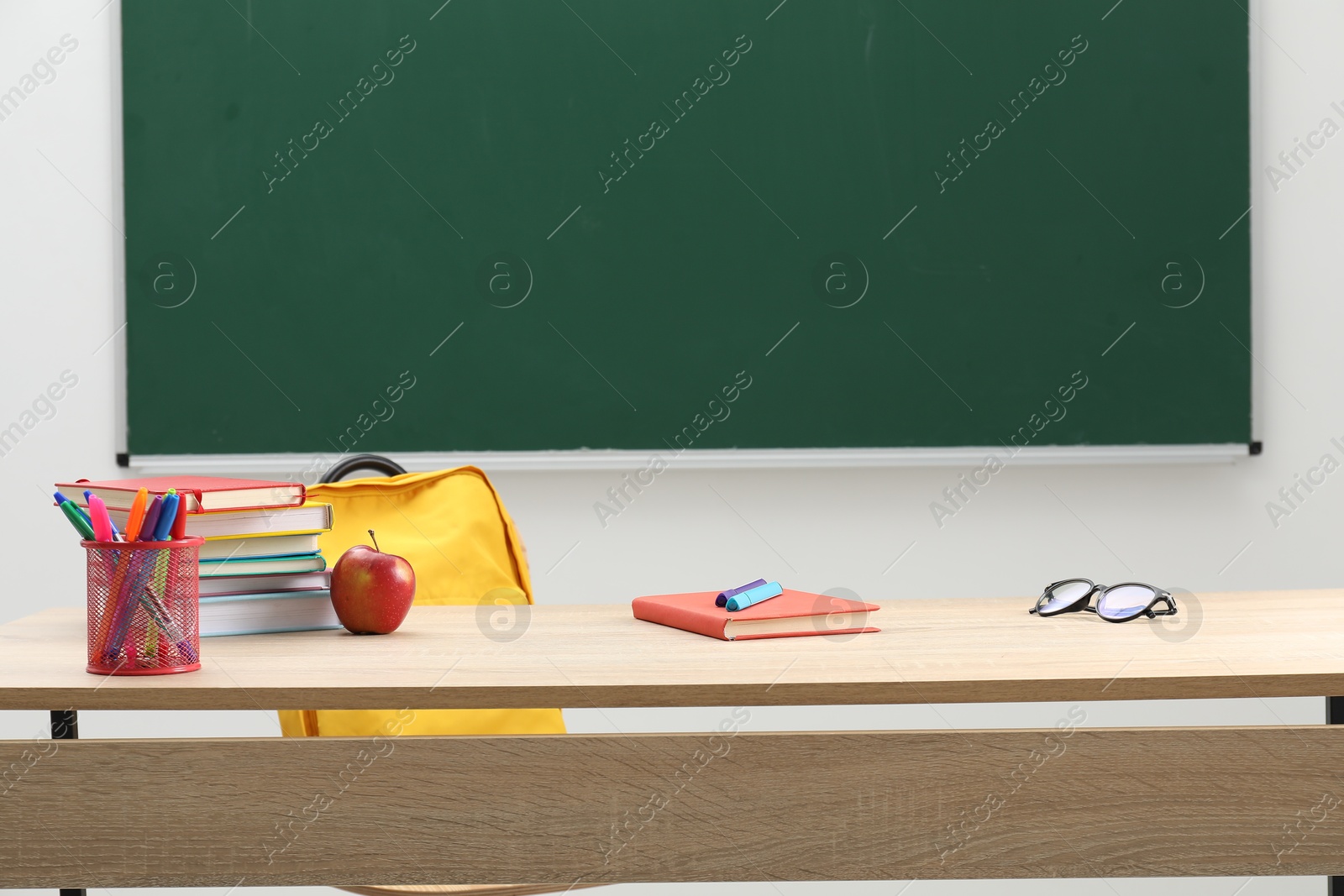 Different school stationery, glasses and red apple on wooden table near blackboard indoors Photo of Different school stationery, glasses and red apple on wooden table near blackboard indoors