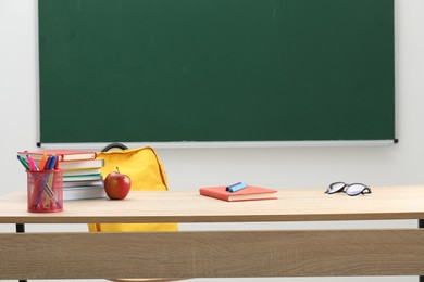 Photo of Different school stationery, glasses and red apple on wooden table near blackboard indoors