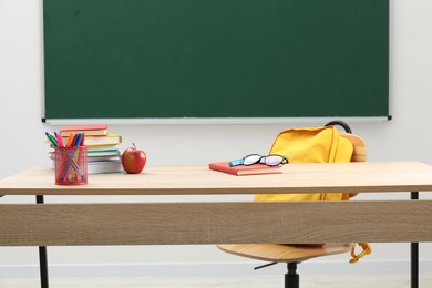 Photo of Different school stationery, glasses and red apple on wooden table near blackboard indoors