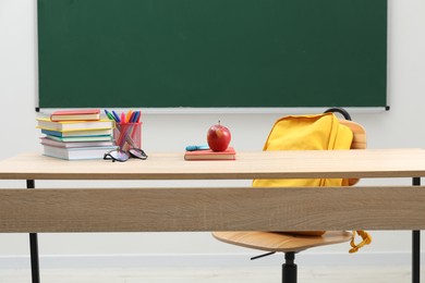 Photo of Different school stationery, glasses and red apple on wooden table near blackboard indoors