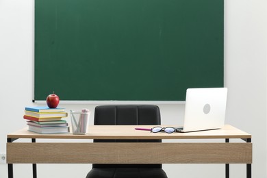 Photo of Different school stationery, laptop, glasses and red apple on wooden table near blackboard indoors