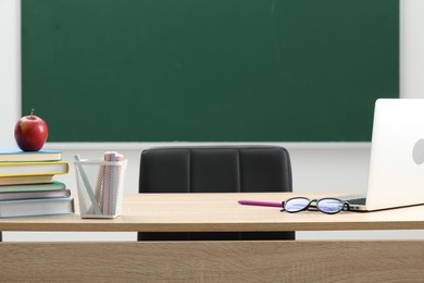 Photo of Different school stationery, laptop, glasses and red apple on wooden table near blackboard indoors
