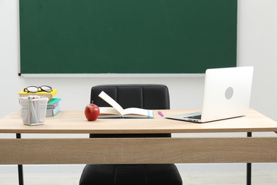 Photo of Different school stationery, laptop, glasses and red apple on wooden table near blackboard indoors