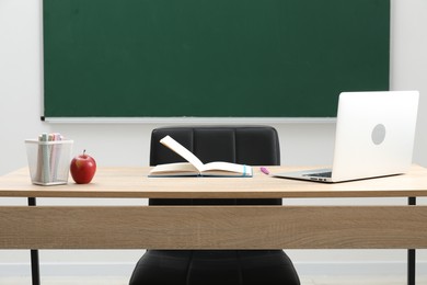Photo of Different school stationery, laptop and red apple on wooden table near blackboard indoors
