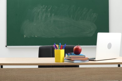 Photo of Different school stationery, laptop and red apple on wooden table near blackboard indoors