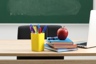 Photo of Different school stationery, laptop and red apple on wooden table near blackboard indoors