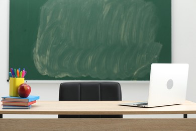 Photo of Different school stationery, laptop and red apple on wooden table near blackboard indoors