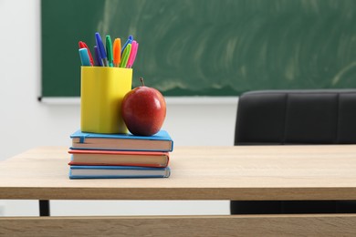 Photo of Different school stationery and red apple on wooden table near blackboard indoors, space for text