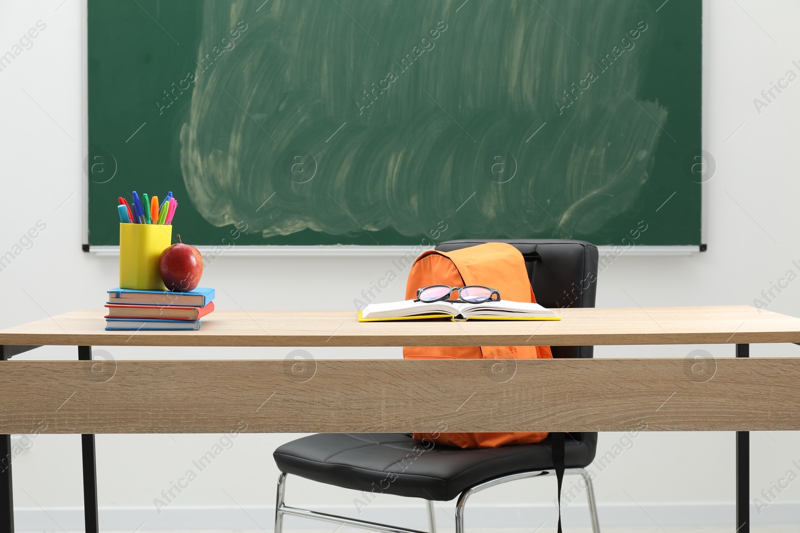 Different school stationery, glasses and red apple on wooden table near blackboard indoors Photo of Different school stationery, glasses and red apple on wooden table near blackboard indoors