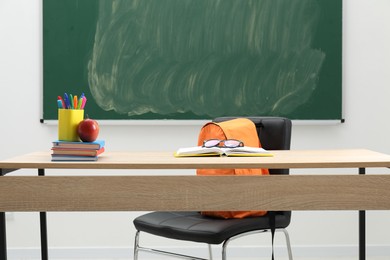 Photo of Different school stationery, glasses and red apple on wooden table near blackboard indoors