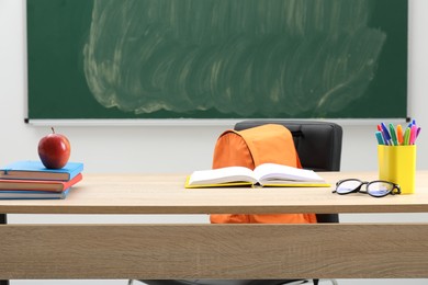 Photo of Different school stationery, glasses and red apple on wooden table near blackboard indoors