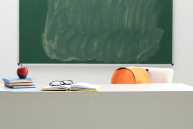 Photo of Different school stationery, glasses and red apple on white table near blackboard indoors