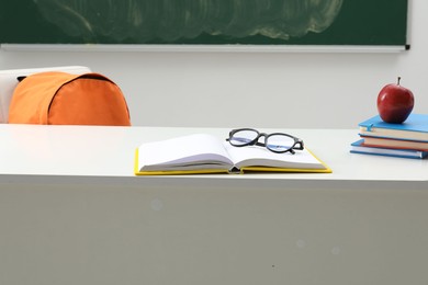 Photo of Different school stationery, glasses and red apple on white table near blackboard indoors