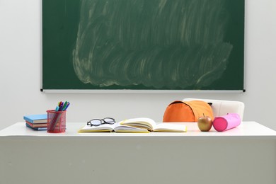 Photo of Different school stationery, glasses and apple on white table near blackboard indoors