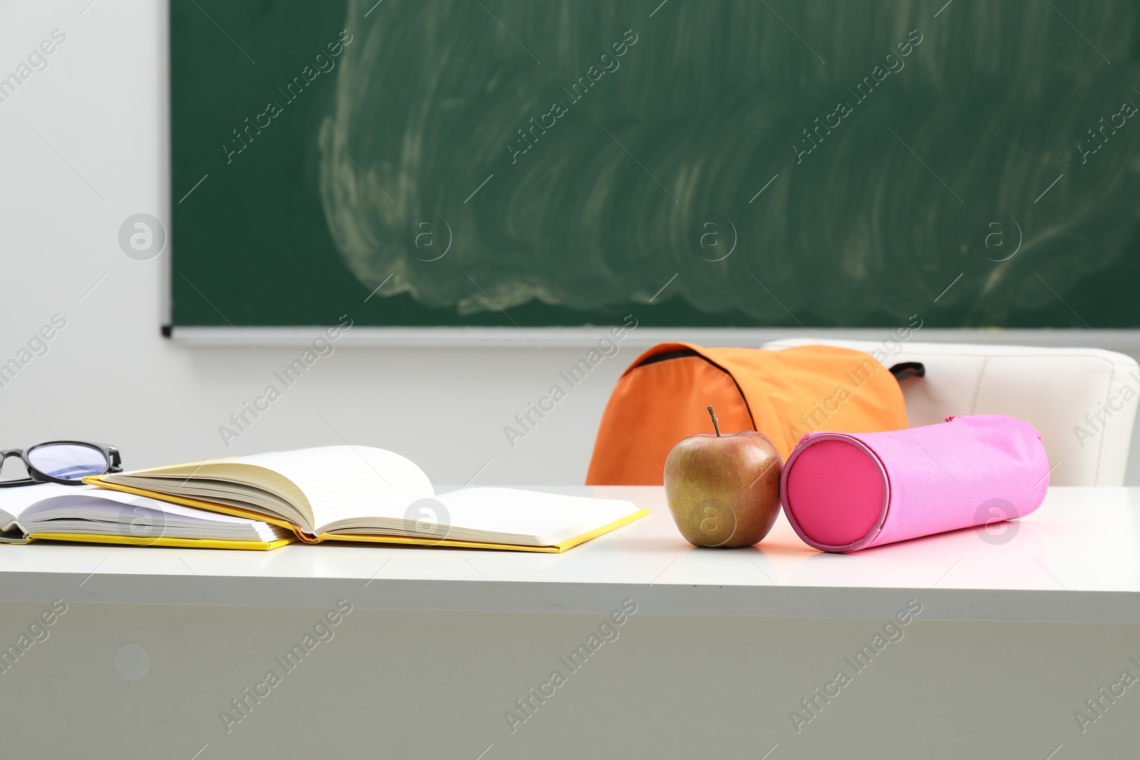 Different school stationery, glasses and apple on white table near blackboard indoors Photo of Different school stationery, glasses and apple on white table near blackboard indoors