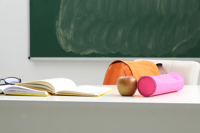 Photo of Different school stationery, glasses and apple on white table near blackboard indoors