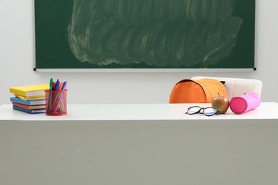 Photo of Different school stationery, glasses and apple on white table near blackboard indoors