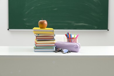 Photo of Different school stationery, glasses and apple on white table near blackboard indoors