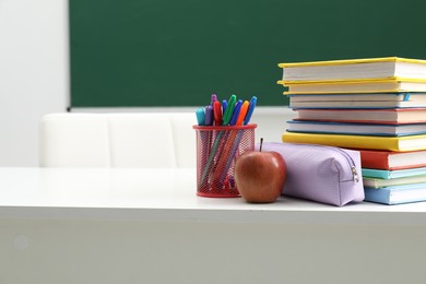 Photo of Different school stationery and apple on white table near blackboard indoors, space for text