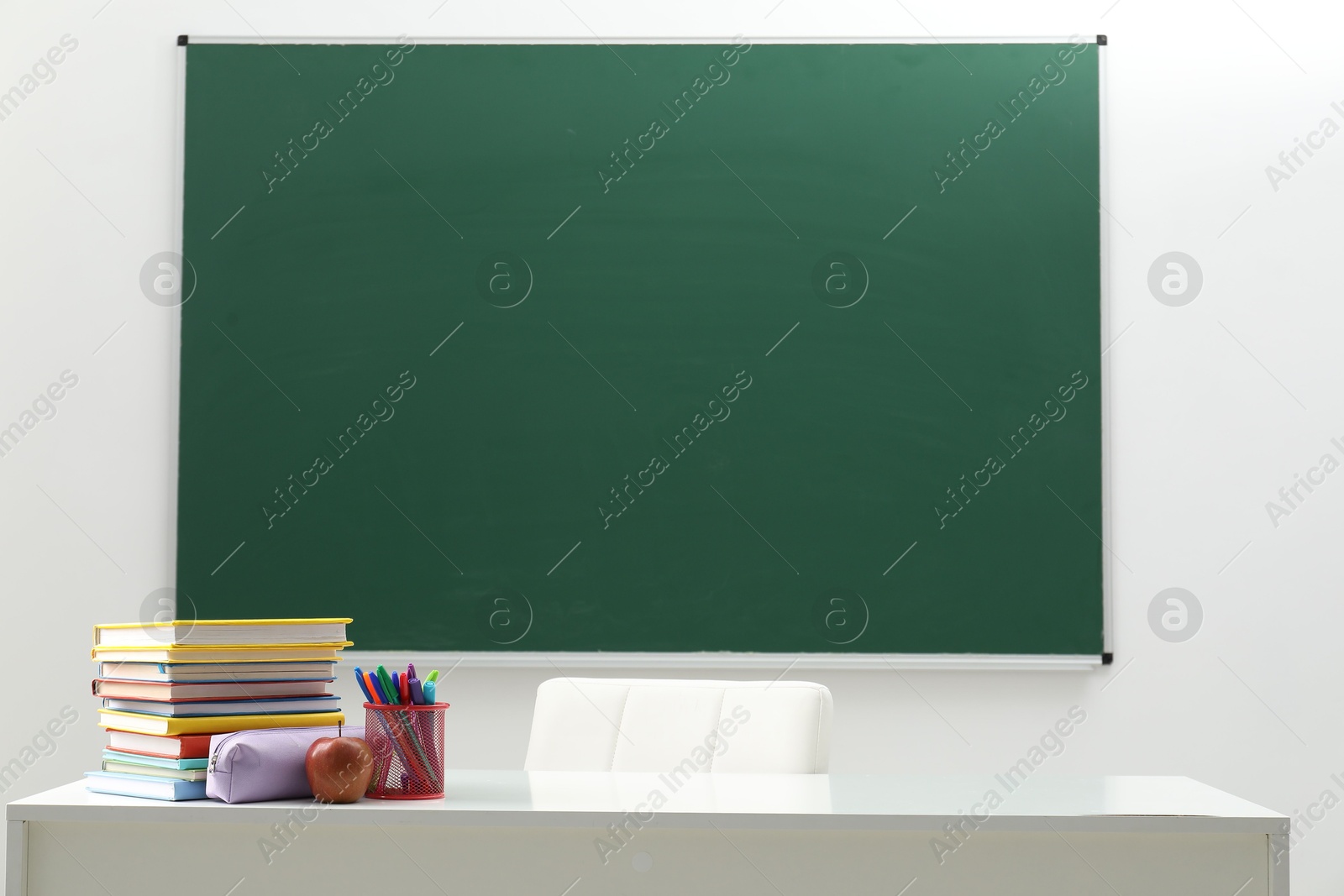 Different school stationery and apple on white table near blackboard indoors Photo of Different school stationery and apple on white table near blackboard indoors