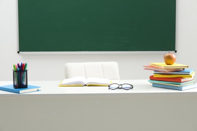 Photo of Different school stationery, glasses and apple on white table near blackboard indoors